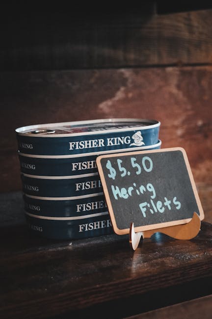 A close-up view of a stack of four round tins labeled 'Fisher King,' placed on a wooden surface with a dark wood-paneled background. In front of the tins, there is a small outdoor chalkboard sign shaped like a fish, displaying the handwritten price of $5.50 and the text 'Haring Fillets,' indicating a sale of fresh fish fillets. The scene suggests a market or outdoor shop setting, with natural lighting highlighting the textures of the wood, tins, and chalkboard. This setup illustrates outdoor sales of seafood products, possibly related to general outdoor or garden-related activities such as preparing for a picnic or backyard barbecue, aligned with gardening and outdoor living themes.