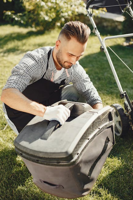 A man with short brown hair and a beard, wearing a checked shirt and black apron, is kneeling on a lush green lawn while servicing a large grey lawnmower. The scene takes place outdoors in a garden with bright, natural sunlight illuminating the area. The garden features well-maintained grass, flower beds, and a pathway with gravel or paving stones. In the background, there are trees and shrubs, and part of a garden bench or raised planting bed is visible. The man appears focused on performing garden maintenance, possibly cleaning or adjusting the mower, with other gardening tools or equipment nearby. The setting reflects professional or dedicated lawn care work, aligning with the theme of affordable garden services offered by Gardeners Harringay, as referenced on the webpage about garden maintenance and landscaping work.